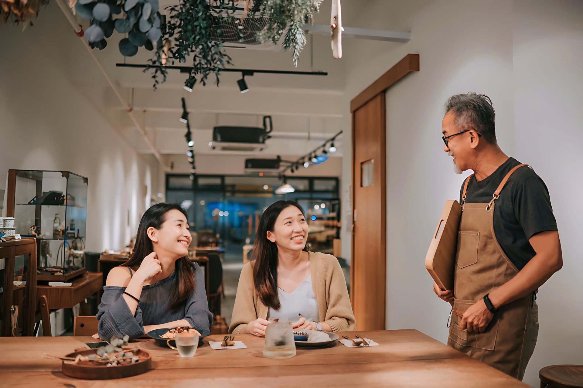 Smiling restaurant owner in an apron talking to two happy customers seated at a wooden table in a warmly lit café.