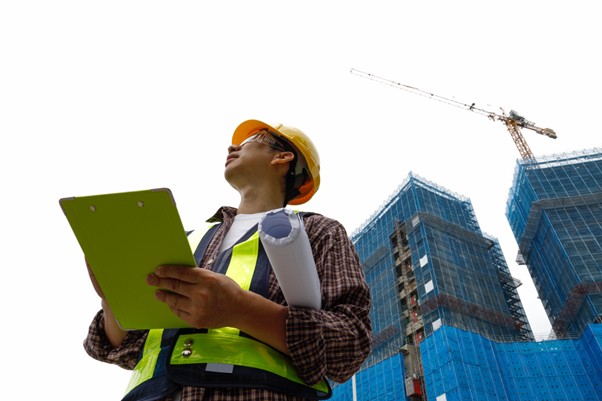 Construction site supervisor in reflective vest and hard hat holding a clipboard and blueprint, looking up at a high-rise building under construction.