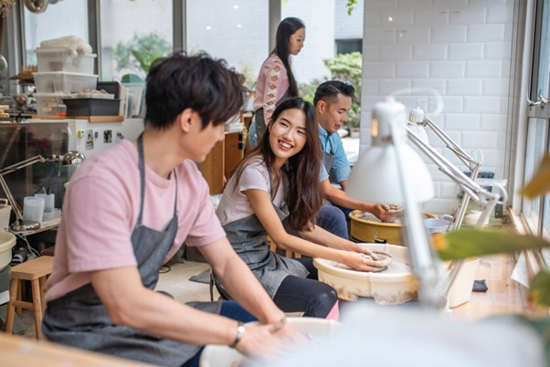 Young adults participating in a pottery class, smiling and engaging with each other while working on clay at a shared studio table.