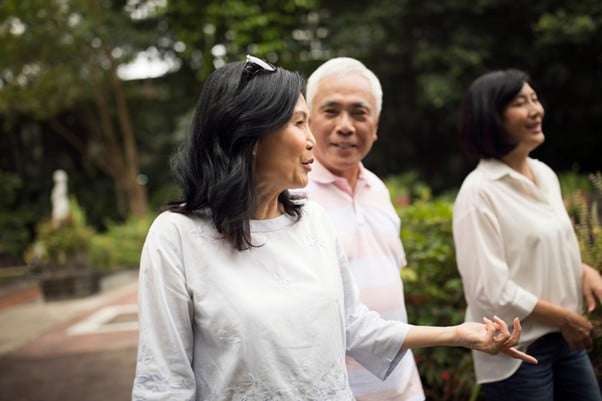 Three older Asian adults walking and chatting together in a peaceful urban park setting, enjoying a relaxed moment outdoors.