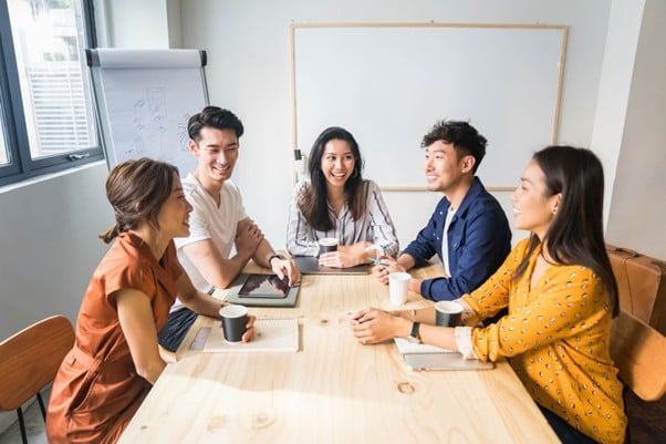 Group of young Asian professionals having a relaxed and cheerful team meeting in a bright modern office, seated around a wooden table with notebooks and coffee cups.