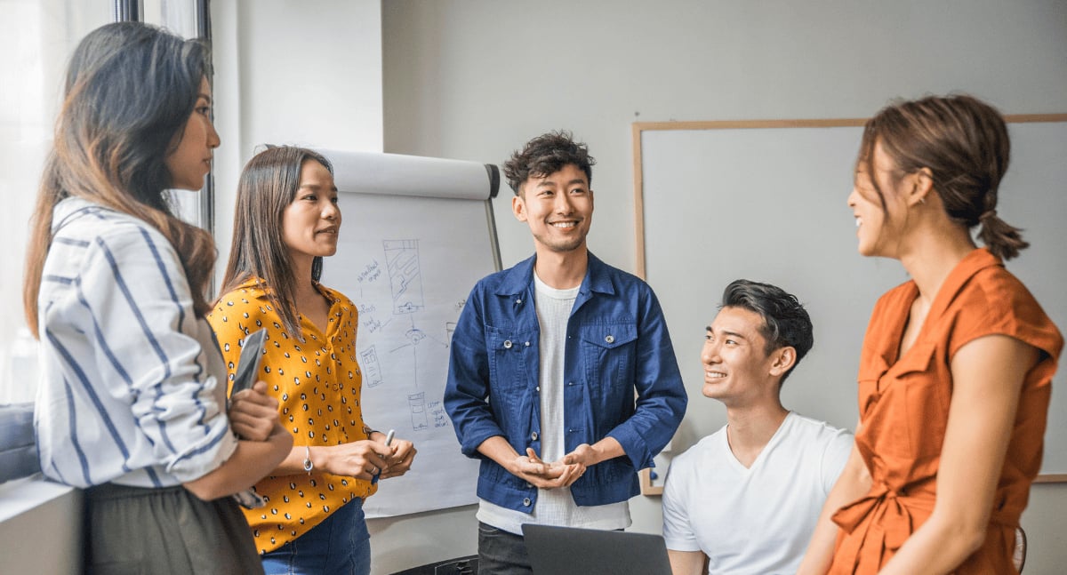 A group of young professionals in a bright Hong Kong office, standing and sitting together, smiling and discussing ideas in front of a flipchart.