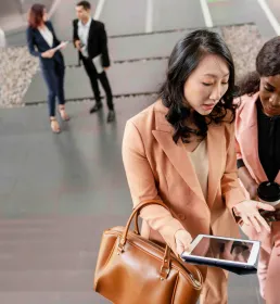 Two professional women in conversation while walking up stairs, one holding a tablet and the other a coffee cup, with colleagues in the background.