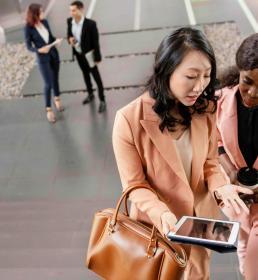 Two professional women in conversation while walking up stairs, one holding a tablet and the other a coffee cup, with colleagues in the background.