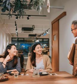 Smiling restaurant owner in an apron talking to two happy customers seated at a wooden table in a warmly lit café.