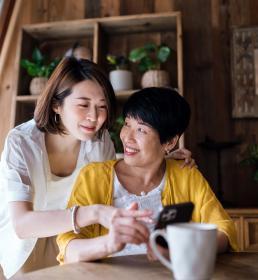 Affectionate Asian senior mother and daughter using smartphone together at home, smiling joyfully, enjoying mother and daughter bonding time. Multi-generation family and technology