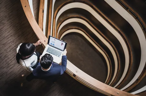 Two finance professionals review accountancy data on a laptop looking out over a spiral staircase