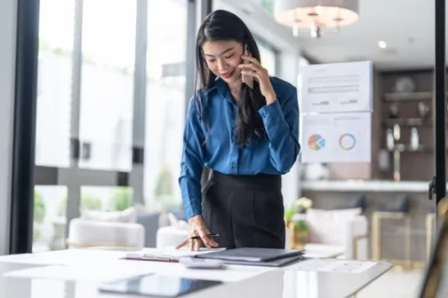 Professional woman standing in a stylish office, talking on the phone and reviewing paperwork