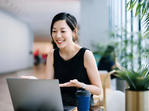 Smiling professional woman sitting at a desk with a laptop and coffee cup, engaged in a video call in a bright, modern indoor setting with natural light and greenery.