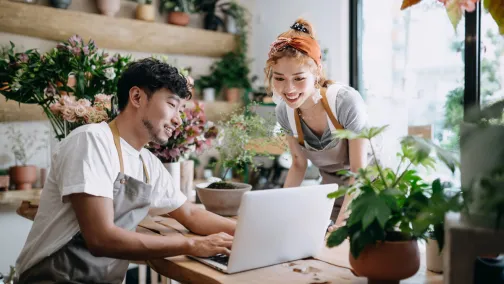 Two young entrepreneurs in aprons smiling while working on a laptop in a flower shop, surrounded by lush plants and floral arrangements.