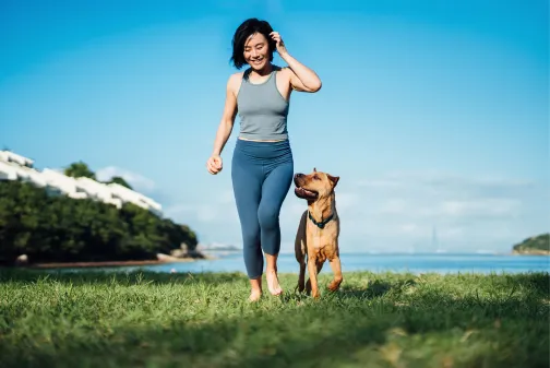 Happy young Asian woman and her pet dog running by the beach against clear blue sky, playing and enjoying time together in the nature outdoors.