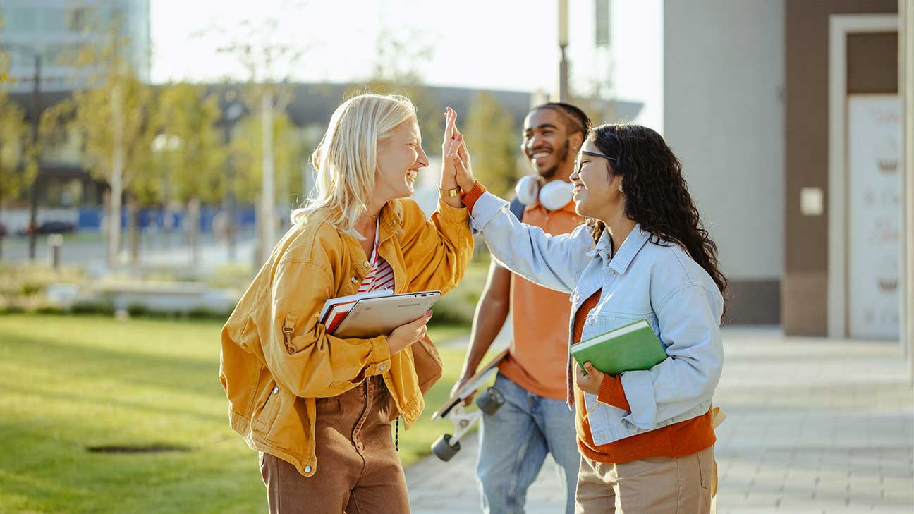 Three people outdoors, two women high-five while holding books, a man smiles in the background with a skateboard.