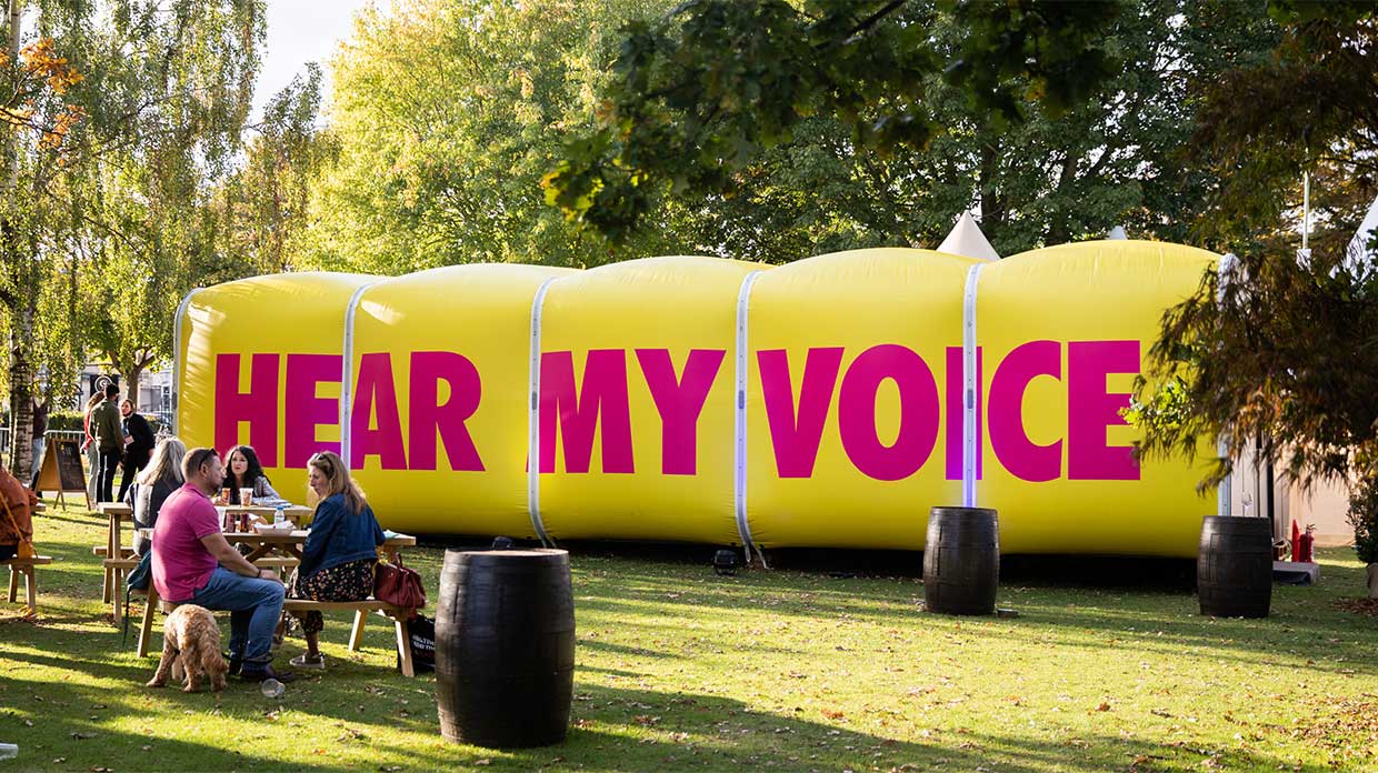 A large yellow inflatable structure with "HEAR MY VOICE" in bold pink letters, surrounded by people on a grassy area.