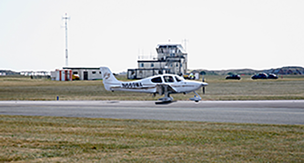 A small white propeller airplane taxis on a runway near a control tower and parked cars on a grassy airfield.