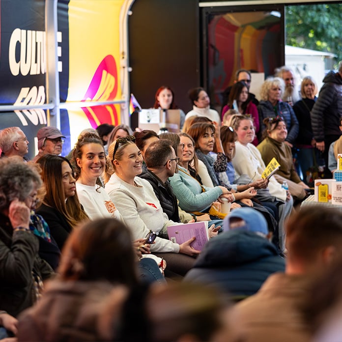 A seated audience listens attentively at an indoor cultural event, with signs and colourful decor in the background.