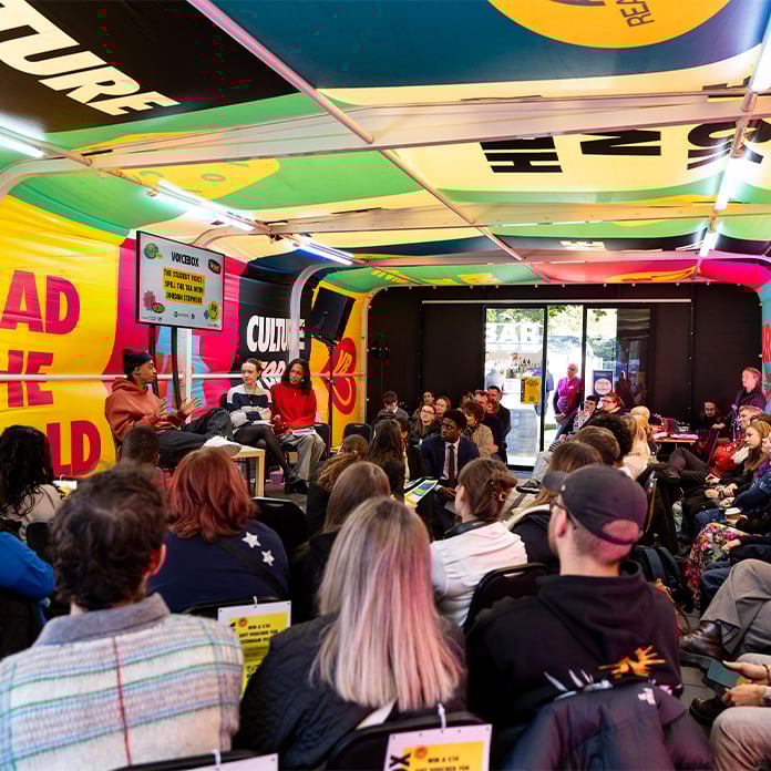 A diverse audience listens to a panel discussion inside a colourful, branded event tent with banners and a presentation screen.