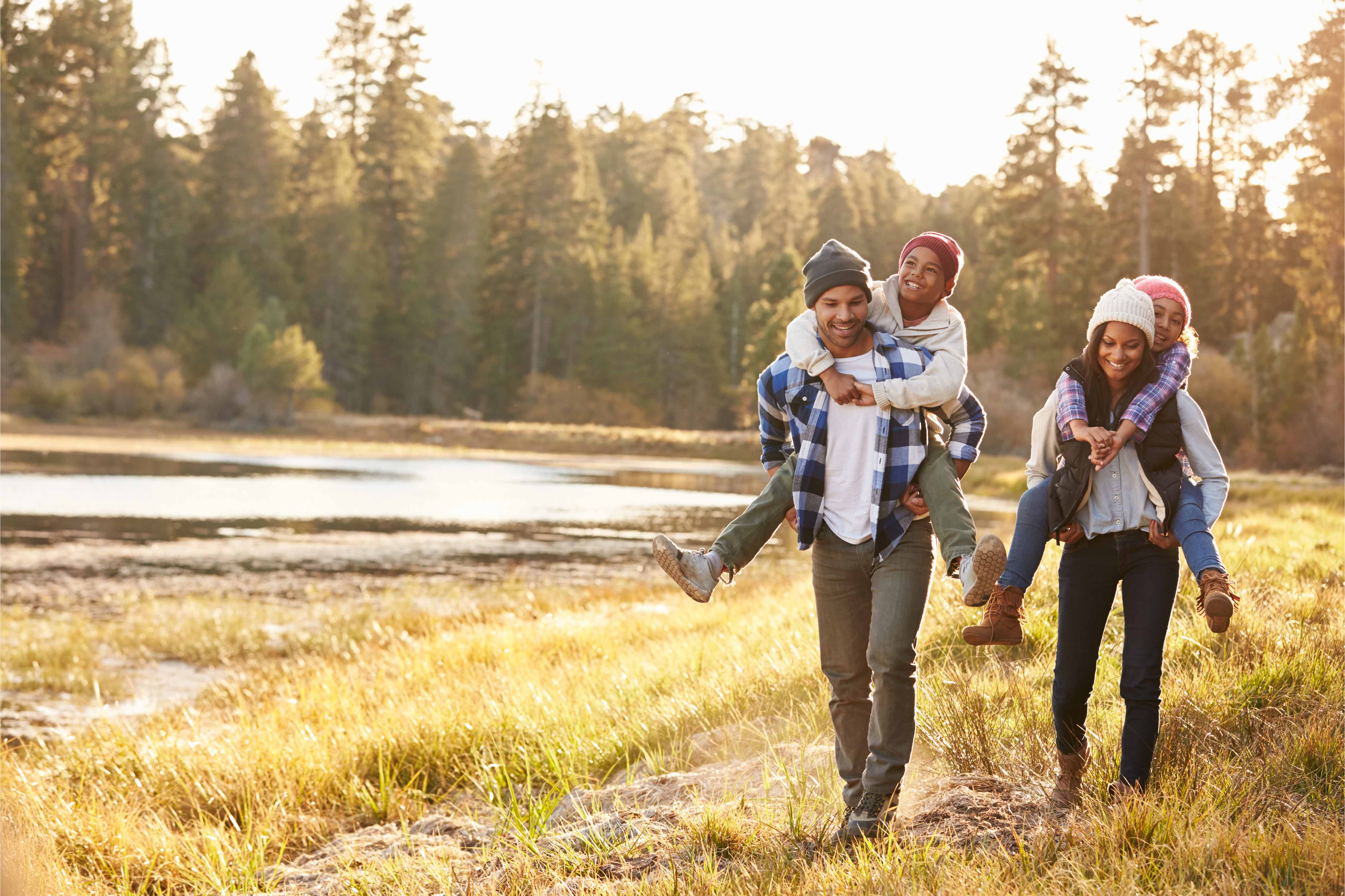 Family of four walking in nature, with parents giving children piggyback rides, smiling and enjoying the outdoors.