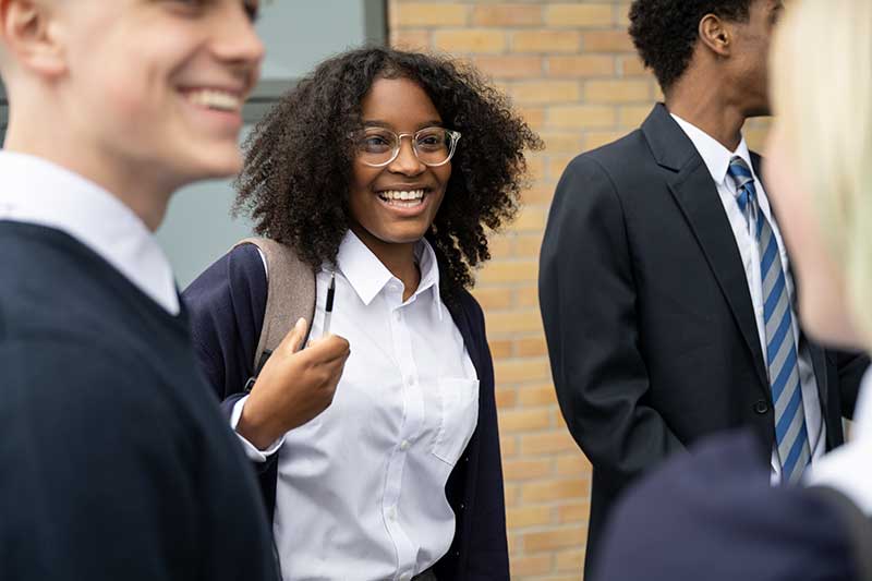 Group of students in school uniforms standing outside, with one student in focus smiling and holding a backpack.