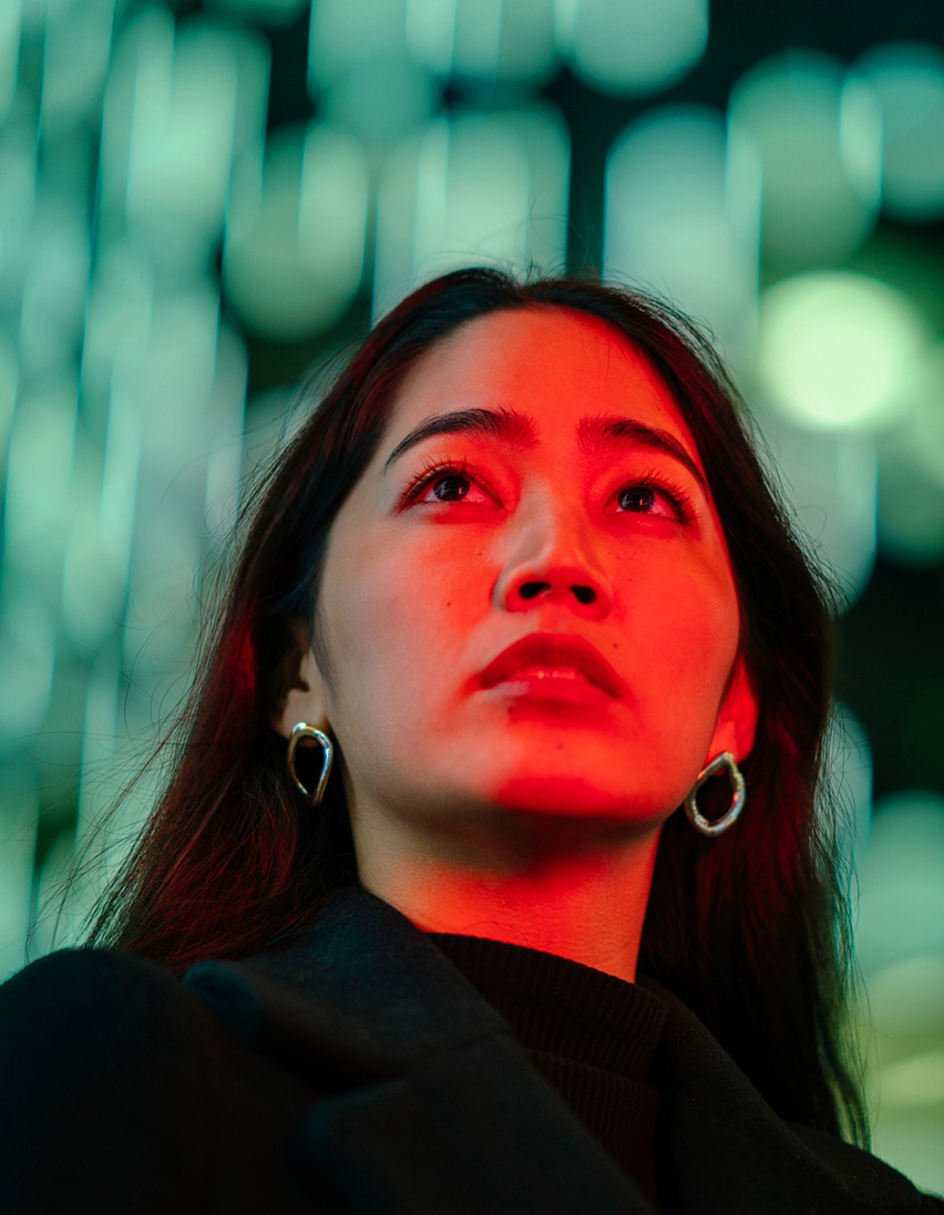 Woman with long dark hair looks upward, her face lit by red light against a blurred background of teal lights.