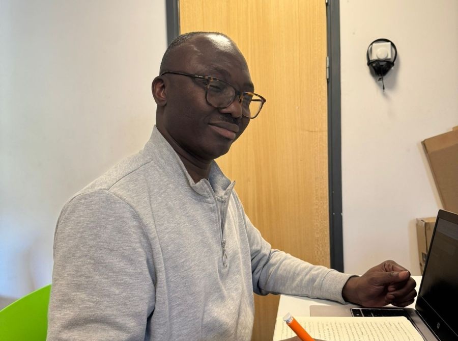 A man wearing glasses sits at a desk, writing in a notebook with an orange pen next to a laptop.