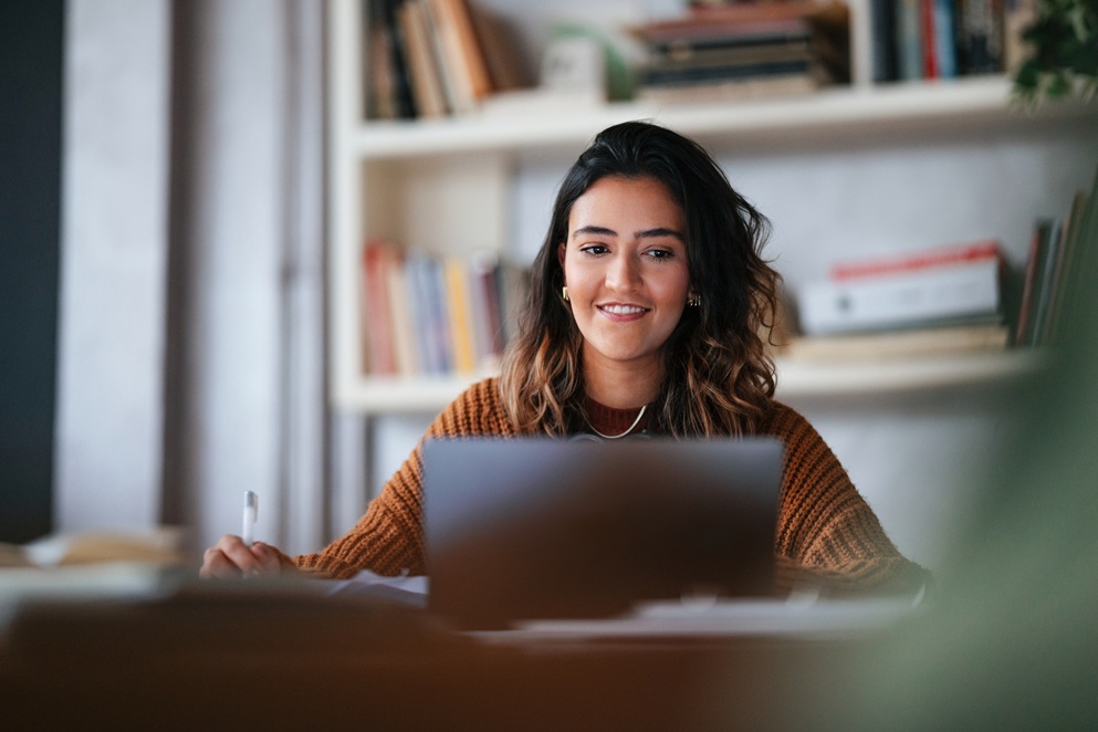 Woman sits at a desk smiling and working on a laptop, holding a pen, with bookshelves in the background.