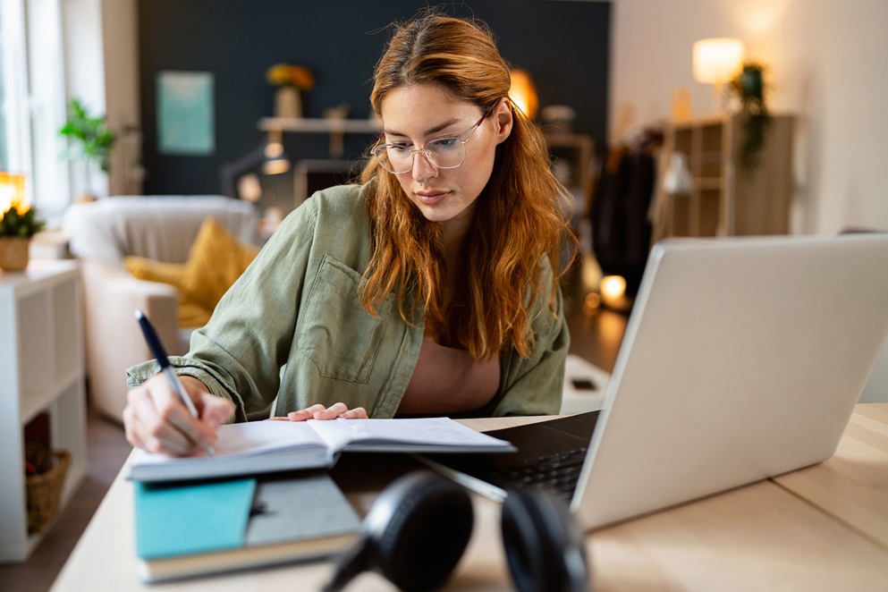 Woman with glasses writing in a notebook at a desk with a laptop and headphones in a home office setting.