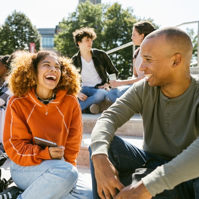 A group of young adults sit on outdoor steps, talking and smiling on a sunny day near a building with trees in the background.