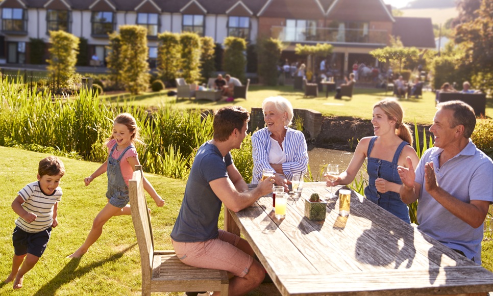 Three adults and two children sit at an outdoor table, while two kids run on grass in a sunny garden near a building.