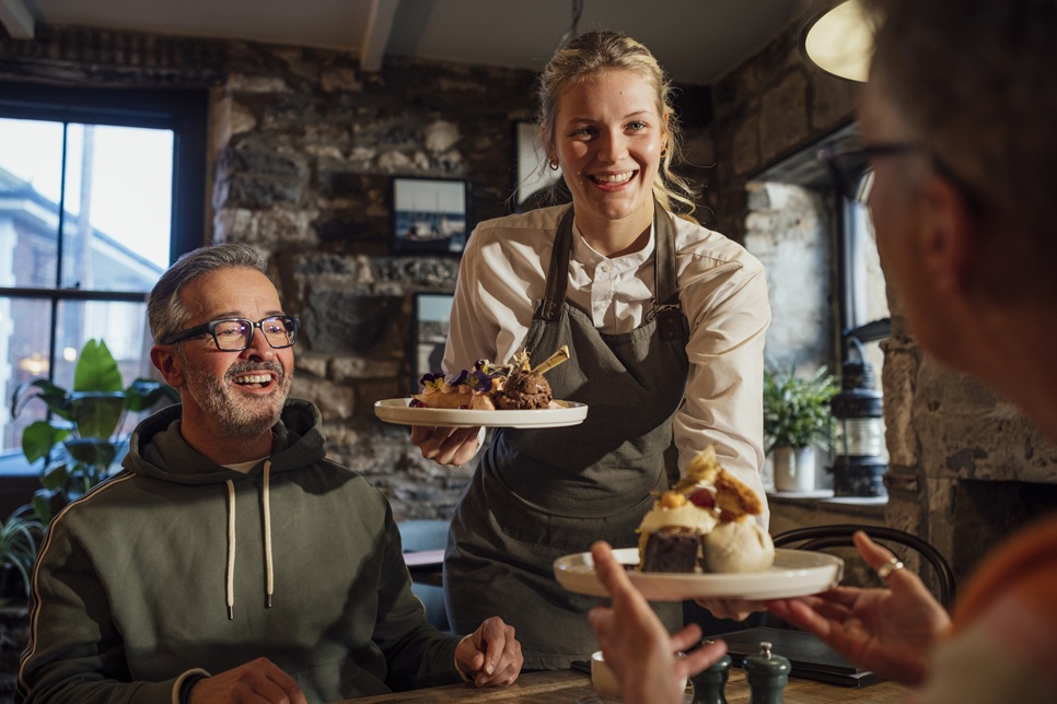 A waitress serves dessert to a smiling seated man in a cozy restaurant with stone walls and natural light.