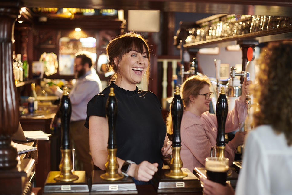 A woman smiles while serving drinks behind the bar in a busy pub, with other staff working in the background.