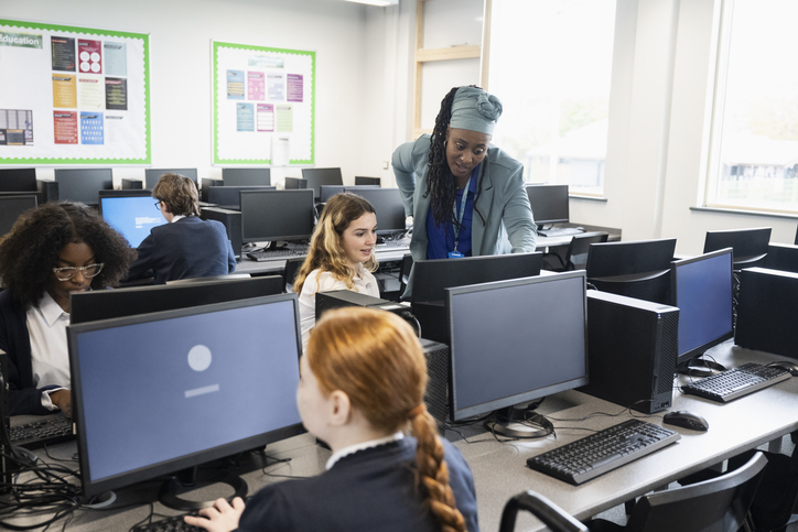 A woman assists students with their work in a computer lab, providing guidance and support at their computer stations.