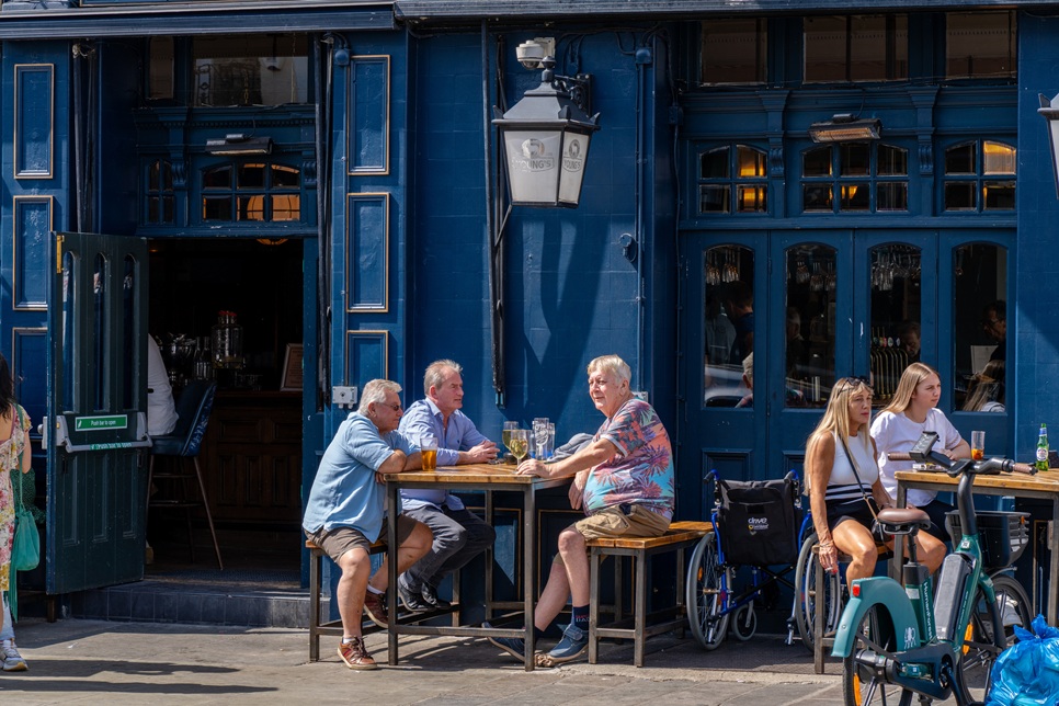 People sit at outdoor tables in front of a blue-painted pub on a sunny day, with drinks and bicycles nearby.