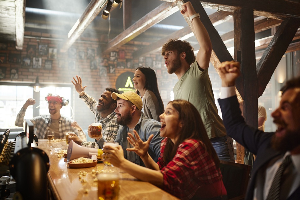 A group of people in a bar cheer and raise their arms while watching an event on a screen, with drinks and snacks on the table.