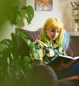 Student sat in a chair reading a book next to a monstera houseplant