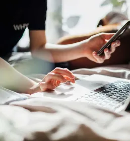 A person holding a phone in one hand and typing on a laptop with the other surrounded by paperwork