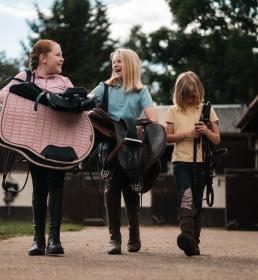 Three girls walking through yard carrying saddles and equipment for tacking up