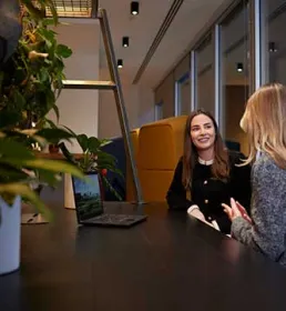Two women sit and talk at a table in a modern office space, with plants and a laptop visible in the foreground.