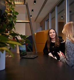 Two women sit and talk at a table in a modern office space, with plants and a laptop visible in the foreground.