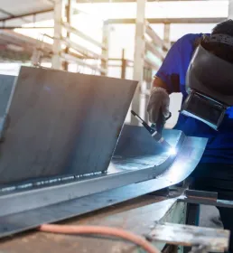 Industrial worker using TIG welding equipment on a curved metal structure in a factory. The welder wears safety gloves and a protective helmet in a workshop setting.