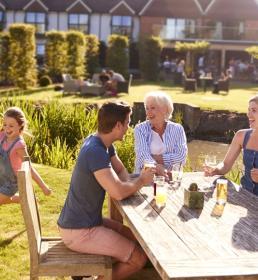 Three adults and two children sit at an outdoor table, while two kids run on grass in a sunny garden near a building.