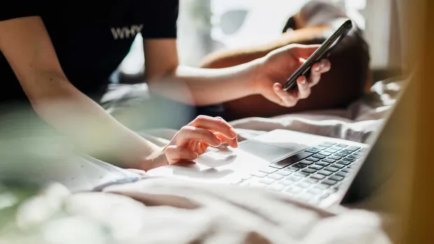 A person holding a phone in one hand and typing on a laptop with the other surrounded by paperwork