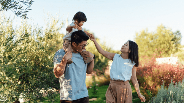 A man outside with child on his shoulders and a woman next to them holding hands with the child