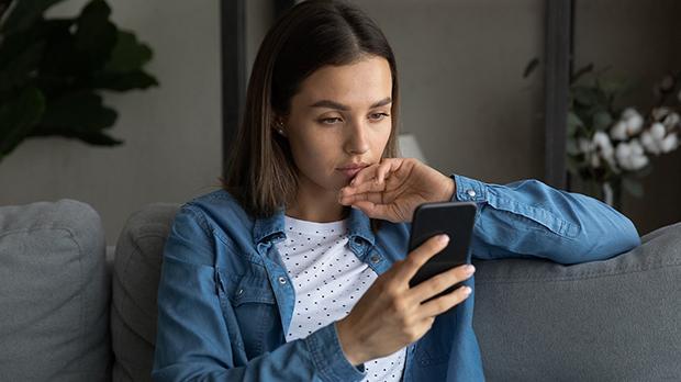 A woman sits on a couch, looking thoughtfully at her smartphone while resting her hand near her mouth.