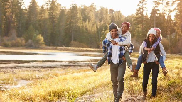 Family of four walking in nature, with parents giving children piggyback rides, smiling and enjoying the outdoors.