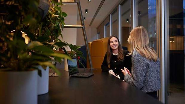Two women sit and talk at a table in a modern office space, with plants and a laptop visible in the foreground.