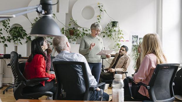 Five people in an office sit in a casual meeting, one woman standing and speaking while others listen attentively.