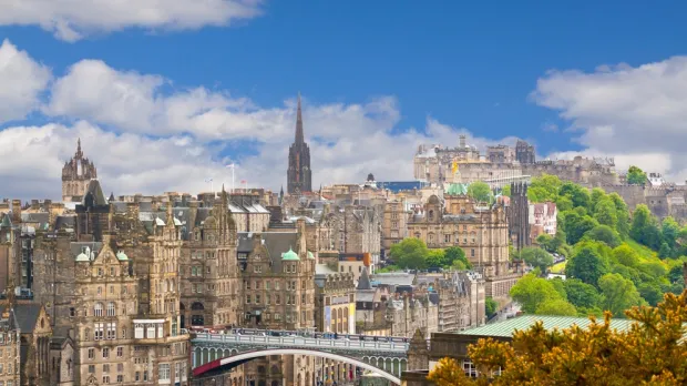 Edinburgh cityscape with historic stone buildings, a bridge, and Edinburgh Castle on a hill under a partly cloudy sky.