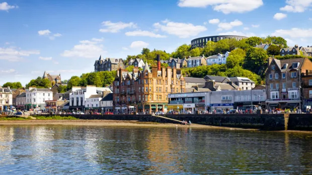 A coastal town with historic buildings, a waterfront, and a circular stone structure on a hill under a blue sky.