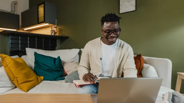 Man sitting on a couch with colourful pillows, using a laptop and writing in a notebook in a living room. Man sitting on a couch with colourful pillows, using a laptop and writing in a notebook in a living room.