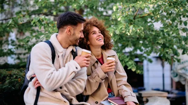 Two people sit outdoors, holding coffee cups and smiling, with books and greenery in the background.